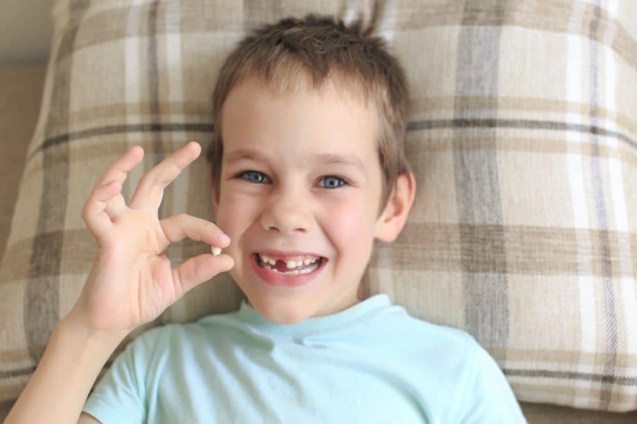 A young boy lying in bed wearing a turquoise shirt is missing his front tooth & smiling while holding a tooth in his hand.