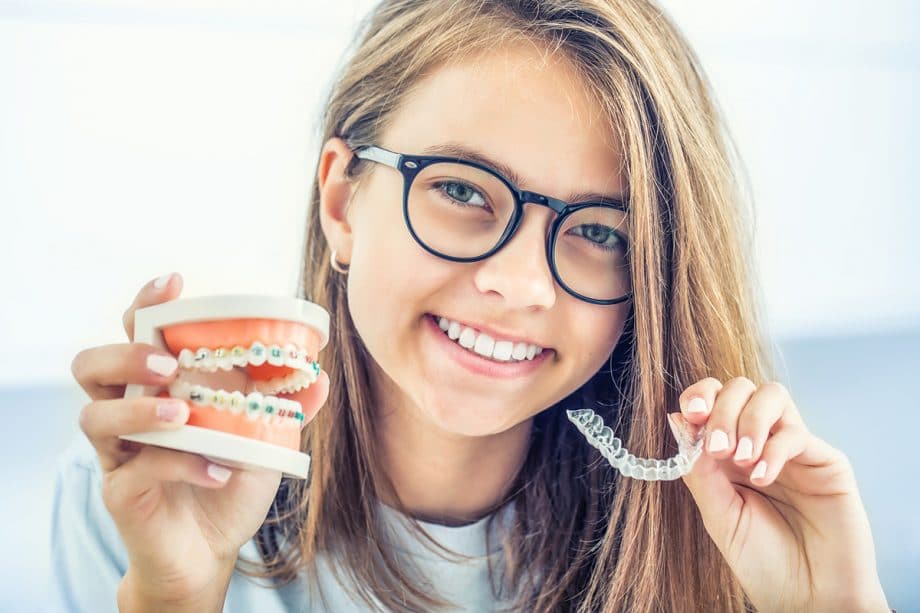 a girl smiles while holding a dental model and an orthodontic retainer